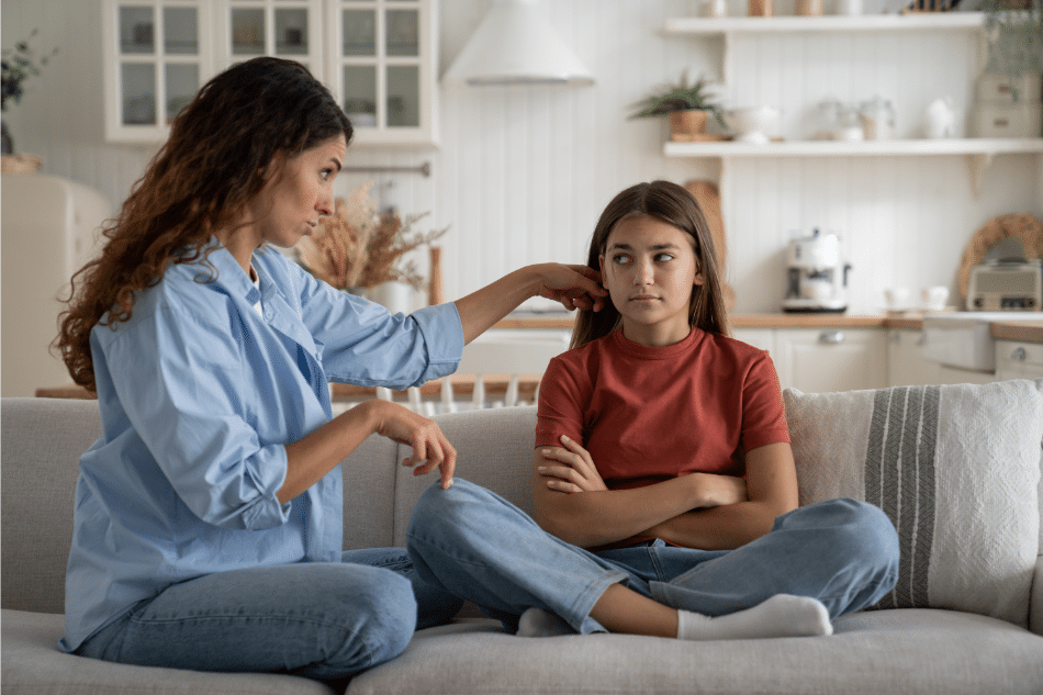 Angry teenage girl sitting cross-legged on the couch while mom tries to console her