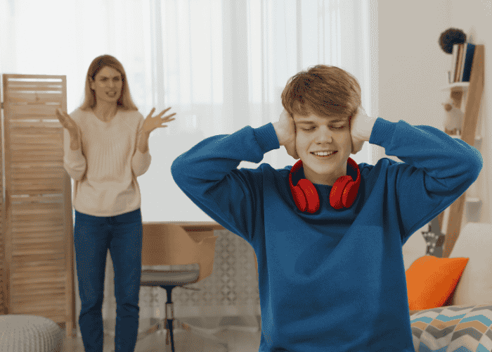 teenage boy with emotional & behavioral symptoms in teens with his hands over his ears while his mum looks at him, angry