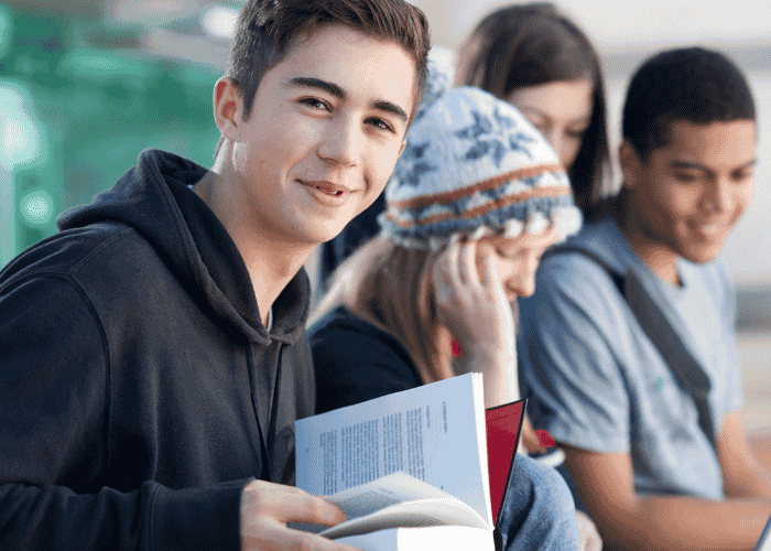 teenage boy holding a book sitting outside with a group of friends smiling after support for people-pleasing in teens