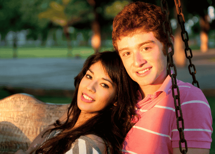 teen boy sitting on swing with girl in park with playing field in background smiling after receiving support for teen codependency