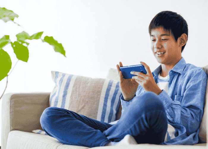 teenage boy sitting cross-legged on sofa playing on phone smiling after receiving treatment for muscle tension and trembling in teens