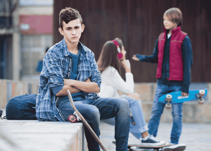 teenage boy at skatepark sitting with arms crossed experiencing teen jealousy & insecurity while another teen boy talks to a teen girl in the background