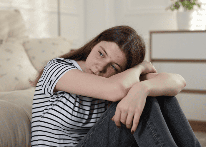 Teenage girl sitting by sofa resting head on arms after self-destructive behavior in teens