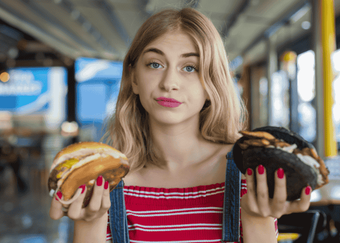 Teenage girl holding up a burger in each hand with a confused look on her face due to teen indecisiveness