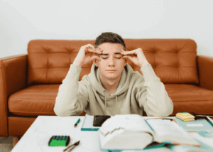 teenage boy sitting at a desk with his eyes closed and fingers on his temples struggling with teen dissociation