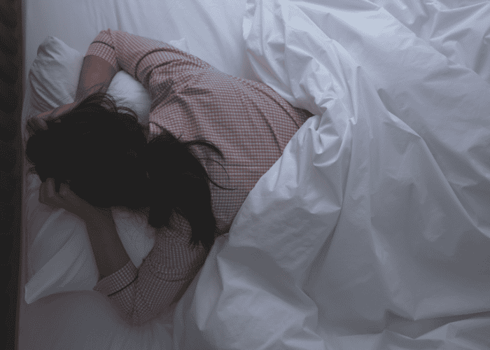 Teenage girl in bed trying to sleep on her front but with her arms up by her head due to teen insomnia