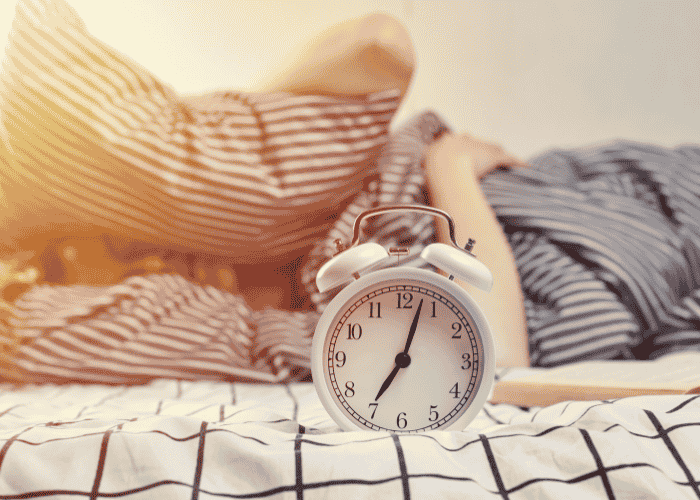 Alarm clock showing 3 minutes past 7 with teenager in the background struggling with oversleeping in teens