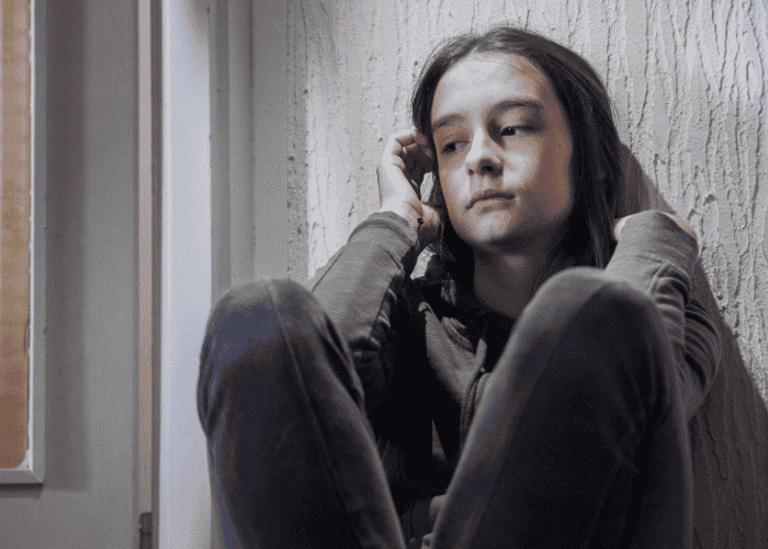 Teenage girl sat on floor with knees pulled to her chest looking upset due to experiencing distressing memories in teens