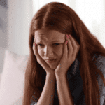 Teenage girl sitting on sofa with head in hands looking anxious experiencing physical symptoms of social anxiety & panic in teens