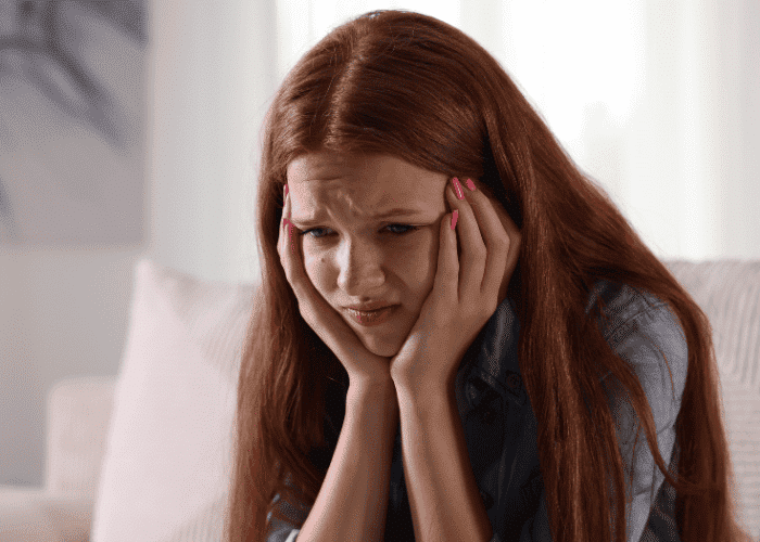 Teenage girl sitting on sofa with head in hands looking anxious experiencing physical symptoms of social anxiety & panic in teens