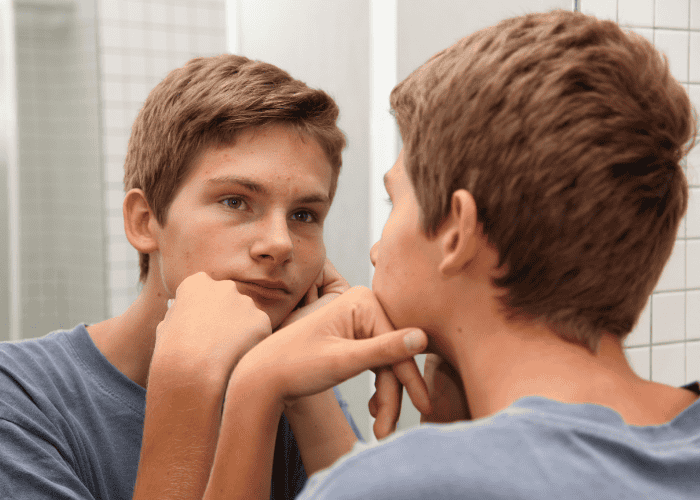 Teenage boy looking in mirror resting chin on hands experiencing harsh self-criticism in teens