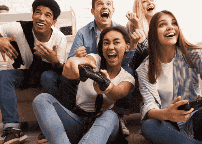 teenage girl sitting cross-legged on the floor playing video games with group of peers smiling after support for avoiding risky decision-making in teens