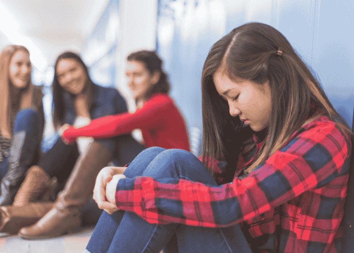 Teenage girl sitting on her own with a group of friends behind her, experiencing fear of being rejection in teens