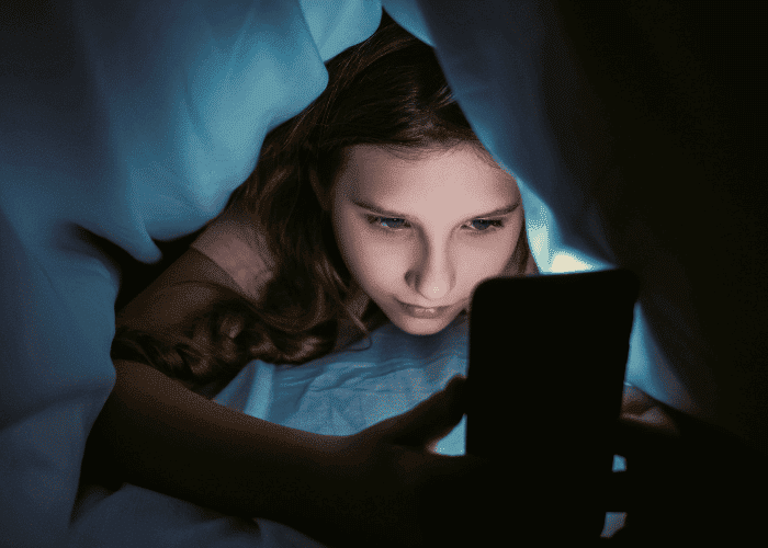 Teenage girl on her phone under the quilt due to irregular or disrupted sleep schedules