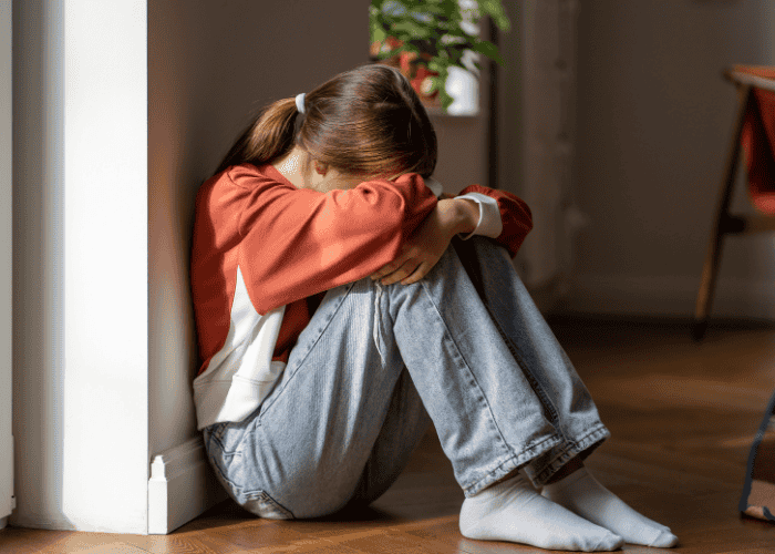 teen girl sat on floor against wall with her knees raised and her face buried in her folded arms needing support from Sharp mental health insurance
