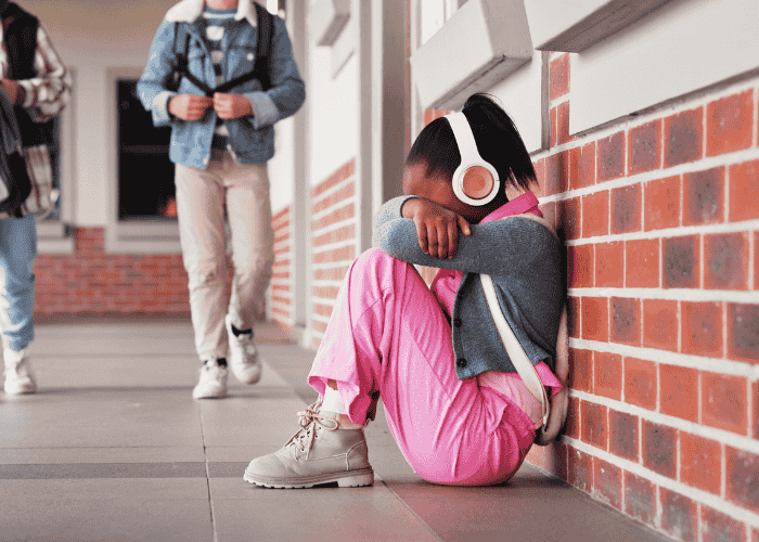 teen girl sitting outside at school against wall with knees pulled to chest and head buried in arms with headphones on wanting help with why sensory overload in teens feels like panic