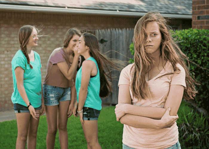 Teen girl experiencing Rejection Sensitive Dysphoria (RSD) in teens standing in garden with arms crossed looking upset as three other girls talk behind her back
