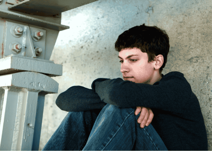 Teen boy sitting outside against wall with knees drawn up to chest and arms resting on knees wondering what Is complex PTSD?
