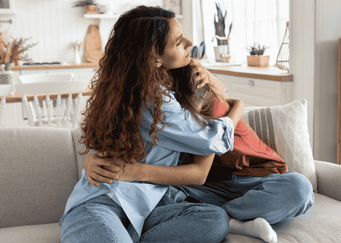 Mother sitting on sofa hugging upset teenage daughter considering mental health insurance cover.