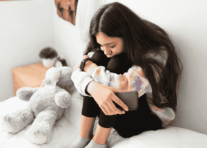 Teenage girl sitting on bed with legs pulled to chest wondering when is the right time to talk to someone about your mental health.