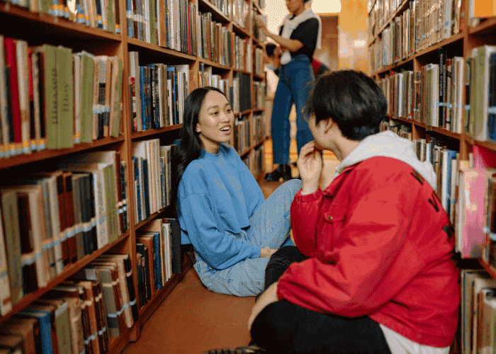 Teenage girl sitting on floor in library chatting to friend about coping with mental health issues