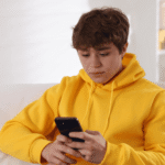 Teenage boy sitting on sofa looking at phone experiencing teen climate anxiety