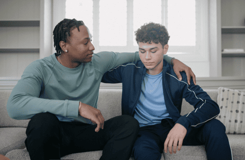 Man with arm around teenage boy sitting together on a couch in a bright interior space.