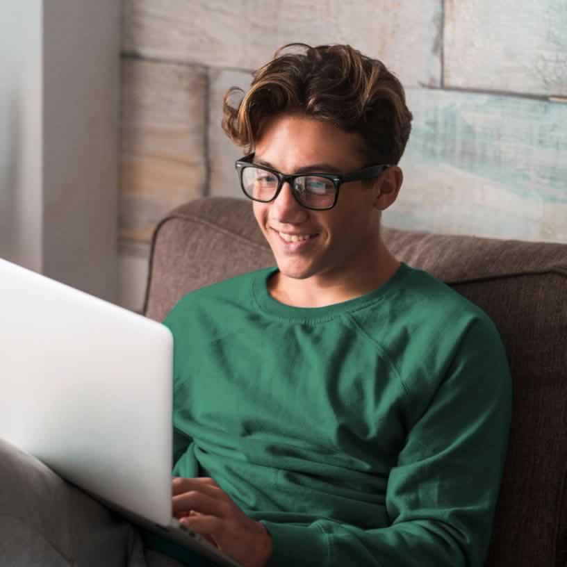 Teen boy sitting on couch with laptop in front of him smiling at the screen