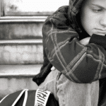 Teen boy sitting on step hunched with chin resting on knees experiencing youth loneliness