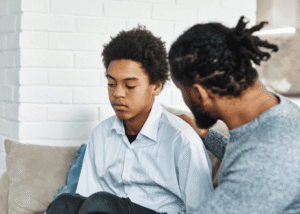 Dad sitting with teenage boy on sofa wondering how to keep communication open with a teen about mental health.