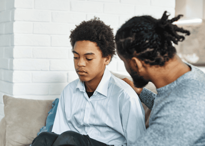 Dad sitting with teenage boy on sofa wondering how to keep communication open with a teen about mental health.