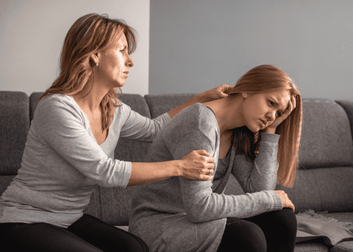Mom sitting on couch with teen daughter with hand on shoulder worrying about distance from home for teen treatment