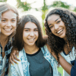 Three teen girls smiling with arms around each others shoulders outside among trees