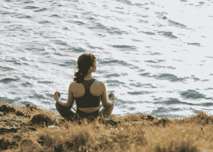Teen girl meditating sitting cross-legged on river bank after support with spiritual identity exploration in teens