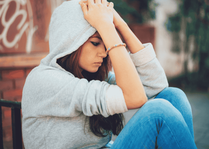 Teen girl sitting outside on bench with hood over head and arms covering face worrying about stigma in teens.