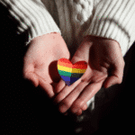 Teen hands holding a rainbow paper heart needing support with self-harm risks in LGBTQ+ teens
