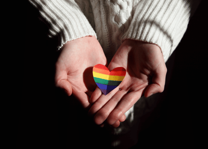 Teen hands holding a rainbow paper heart needing support with self-harm risks in LGBTQ+ teens