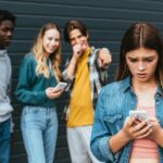 Young teen girl looking at her cell phone while peers stand behind her laughing and pointing at her
