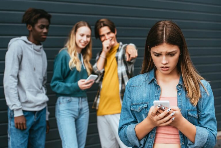 Young teen girl looking at her cell phone while peers stand behind her laughing and pointing at her
