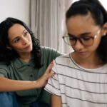 Mom sitting on couch next to daughter while offering support and holding her shoulder