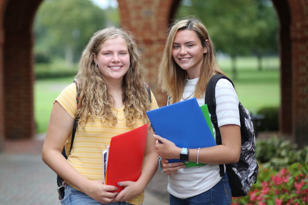 Two teenage girls smiling next to each other wearing backpacks and holding school folders