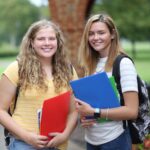 Two teenage girls smiling next to each other wearing backpacks and holding school folders