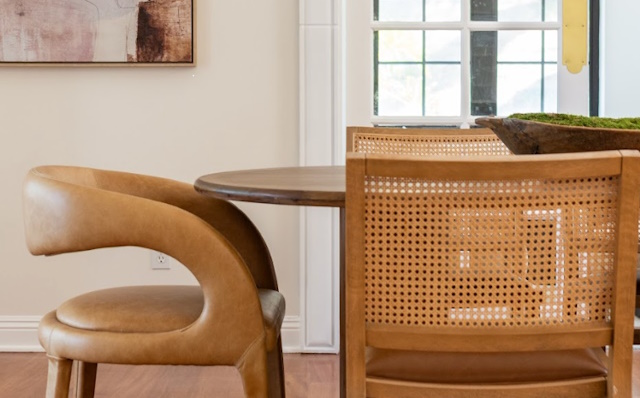 A leather chair and a cane-backed chair beside a round wooden table near a bright window.