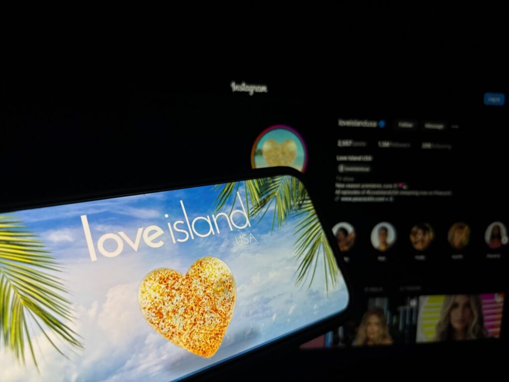 Cell phone screen with Love Island show logo showing on it being held in front of computer screen with Instagram pulled up