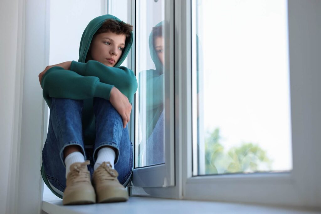 Teen boy in jeans and hoodie sitting on window seat looking sadly out the window