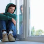 Teen boy in jeans and hoodie sitting on window seat looking sadly out the window