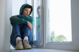 Teen boy in jeans and hoodie sitting on window seat looking sadly out the window
