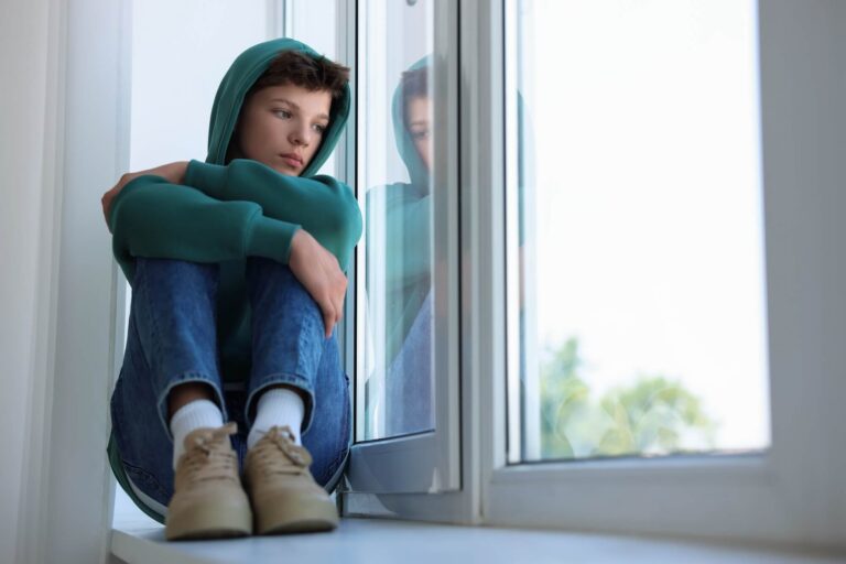 Teen boy in jeans and hoodie sitting on window seat looking sadly out the window