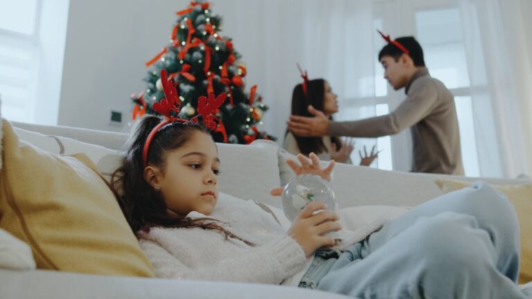 Girl sitting on couch looking at snowglobe while parents fight in the background next to Christmas tree