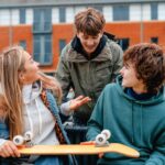 Two teen boys with one teen girl smiling and talking while two of them hold a skateboard